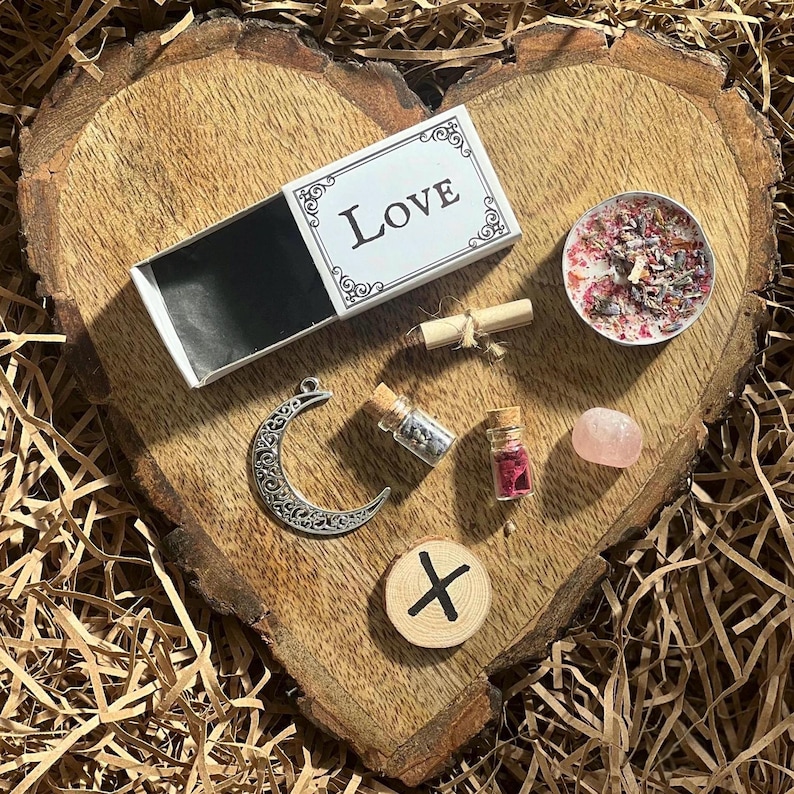 May include: A heart-shaped wooden board with a variety of love-themed items, including a matchbox labeled "Love", a small candle, a crescent moon charm, a small bottle of herbs, a small bottle of crystals, a pink stone, and a wooden rune.
