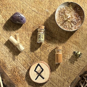 May include: A wooden circle with a black rune symbol, a small glass bottle with a cork stopper filled with dried herbs, a purple amethyst stone, a rolled up piece of paper, a small glass bottle with a cork stopper filled with brown powder, and a small gold bell charm on a wooden surface.