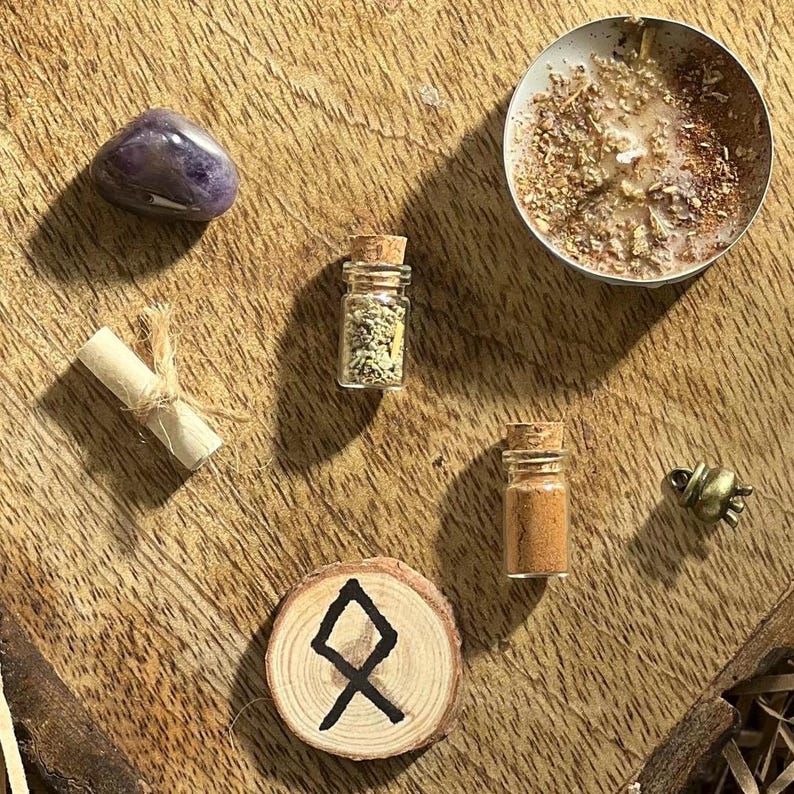 May include: A wooden circle with a black rune symbol, a small glass bottle with a cork stopper filled with dried herbs, a purple amethyst stone, a rolled up piece of paper, a small glass bottle with a cork stopper filled with brown powder, and a small gold bell charm on a wooden surface.