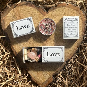 Puede incluir: Una caja blanca con la palabra "LOVE" impresa en ella. La caja contiene una vela pequeña, un cristal de cuarzo rosa, una pequeña botella de aceite y un trozo de papel. La caja está rodeada de virutas de papel marrón.