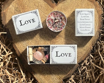 May include: A white box with the word "LOVE" printed on it. The box contains a small candle, a rose quartz crystal, a small bottle of oil, and a piece of paper. The box is surrounded by brown paper shreds.