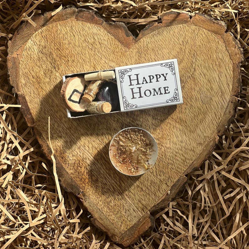 May include: A small white box with the words "Happy Home" on it, a small wooden heart with a symbol on it, and a small candle on a wooden heart-shaped surface.