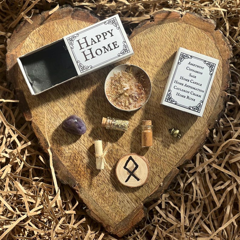 May include: A wooden heart-shaped tray with a variety of items for a home blessing ritual. The items include a small candle, a matchbox labeled "Happy Home", a small bottle of herbs, a small bottle of oil, a small amethyst crystal, a small wooden rune, and a small charm.