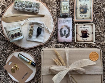 Op de afbeelding: Een verzameling heksen-thema items, waaronder een kaars, orakelkaarten, badzout en een geschenkdoos. De items zijn gerangschikt op stro, met een houten schijf en een gouden veer. De doos is verzegeld met een wasstempel.