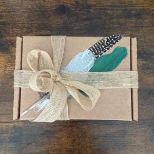 Peut inclure: Une petite bo&icirc;te en carton marron attach&eacute;e avec de la ficelle naturelle et un grand n&oelig;ud. La bo&icirc;te est d&eacute;cor&eacute;e de trois plumes : blanche, verte et &agrave; motifs noirs et blancs. L'emballage rustique sugg&egrave;re un cadeau ou du mat&eacute;riel d'artisanat.