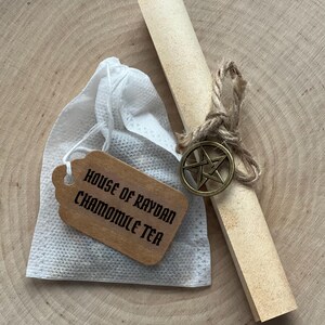 Peut inclure: Un ensemble de th&eacute; &agrave; la camomille. Un sachet de th&eacute; blanc avec une &eacute;tiquette en bois indiquant "HOUSE OF RAYDAN CHAMOMILE TEA". Un parchemin roul&eacute; est attach&eacute; avec de la ficelle et un charme de pentagramme en bronze. Pr&eacute;sent&eacute; sur une surface en bois.