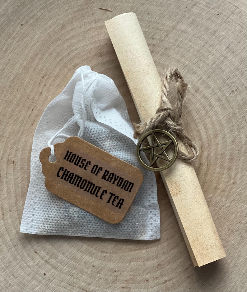 Peut inclure: Un ensemble de th&eacute; &agrave; la camomille. Un sachet de th&eacute; blanc avec une &eacute;tiquette en bois indiquant "HOUSE OF RAYDAN CHAMOMILE TEA". Un parchemin roul&eacute; est attach&eacute; avec de la ficelle et un charme de pentagramme en bronze. Pr&eacute;sent&eacute; sur une surface en bois.