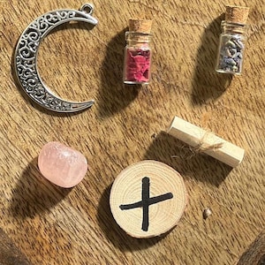 May include: A collection of witchy items including a crescent moon charm, a small wooden disc with a black cross, a pink crystal, a small glass bottle with dried flowers, a rolled up piece of paper tied with twine, and a small glass bottle with dried lavender.