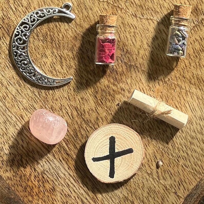 May include: A collection of witchy items including a crescent moon charm, a small wooden disc with a black cross, a pink crystal, a small glass bottle with dried flowers, a rolled up piece of paper tied with twine, and a small glass bottle with dried lavender.