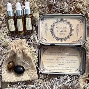 Peut inclure: Un ensemble de trois petites bouteilles en verre avec des bouchons en liège, étiquetées "Protection", "Positivité" et "Mise à la terre". Les bouteilles sont dans une boîte en argent avec le texte "House of Raydan Witches Travel Oils". La boîte comprend également des descriptions des utilisations des huiles. Un petit sac en toile de jute avec trois cristaux est également inclus.