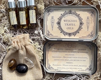 May include: A set of three small glass bottles with cork stoppers, labeled "Protection", "Positivity", and "Grounding". The bottles are in a silver tin with the text "House of Raydan Witches Travel Oils". The tin also includes descriptions of the oils' uses. A small burlap bag with three crystals is also included.