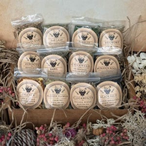 May include: A wooden box filled with small, clear plastic bags containing dried herbs and spices. Each bag has a round label with text such as "St. John's Wort," "Bay Leaf," and "Parsley." The labels feature a mortar and pestle design.