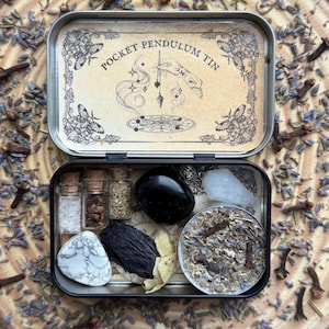 May include: An open, rectangular metal tin labeled "Pocket Pendulum Tin" with a variety of small items inside. The tin contains small glass bottles, crystals, and dried herbs. The background is scattered with dried lavender and cloves.