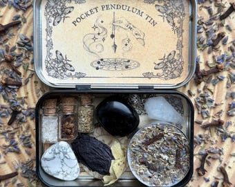 May include: An open, rectangular metal tin labeled "Pocket Pendulum Tin" with a variety of small items inside. The tin contains small glass bottles, crystals, and dried herbs. The background is scattered with dried lavender and cloves.