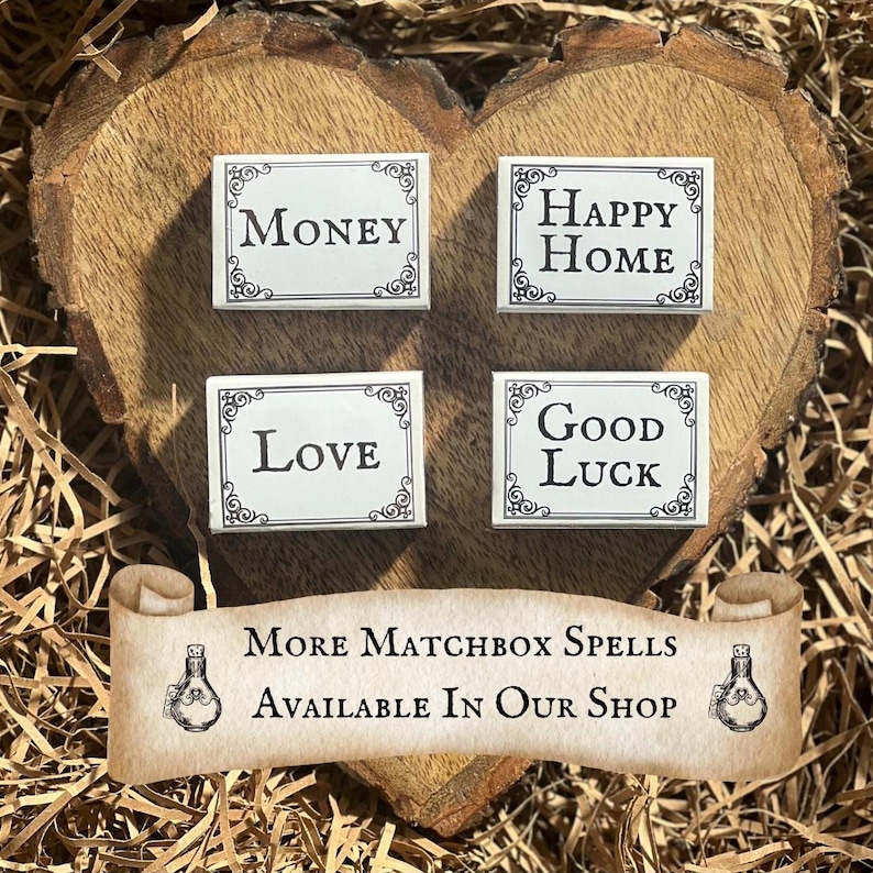 May include: Four white matchboxes with black lettering on a wooden heart-shaped surface. The matchboxes read "Money", "Happy Home", "Love", and "Good Luck". The text "More Matchbox Spells Available In Our Shop" is written on a scroll with a bottle design on each end.