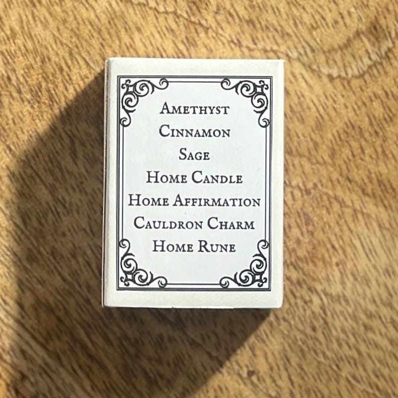 May include: A white box with black text and a decorative border. The text reads "Amethyst Cinnamon Sage Home Candle Home Affirmation Cauldron Charm Home Rune".