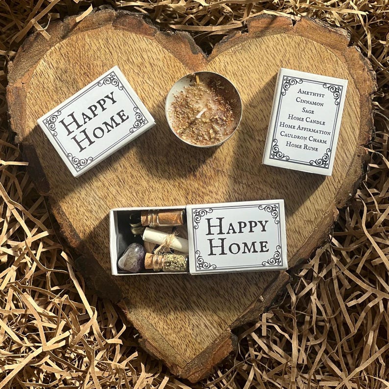 May include: Three white boxes with the words "Happy Home" on them. The boxes contain a candle, a small bottle with a crystal, and a small bottle with herbs. The boxes are on a wooden surface.