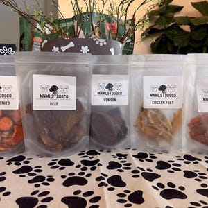 May include: Five clear bags of dog treats are arranged on a paw print tablecloth. The bags contain sweet potato, beef, venison, chicken feet, and chicken. Each bag has a white label with the text "MNMLSTDOOGCO".