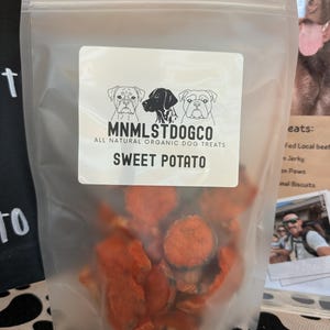 May include: A transparent bag containing orange sweet potato dog treats. The label reads "MNMLSTDOGCO ALL NATURAL ORGANIC DOG TREATS SWEET POTATO" and features illustrations of three dogs. The treats are visible inside.