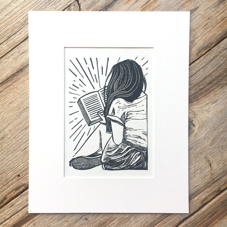 Puede incluir: Grabado en lin&oacute;leo en blanco y negro de una persona sentada leyendo un libro. La persona tiene el pelo largo y lleva una camisa sencilla. El libro est&aacute; abierto y las p&aacute;ginas son visibles. El fondo es un dise&ntilde;o simple de l&iacute;neas radiantes.