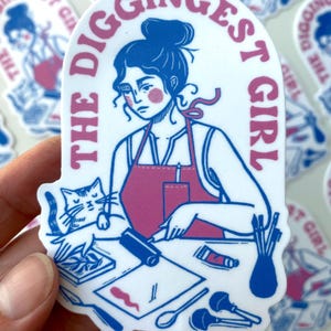 May include: A white sticker with a blue and pink illustration of a woman with a cat, art supplies, and the text "THE DIGGINGEST GIRL". The woman wears a pink apron and has her hair in a bun. The sticker is held in a hand.