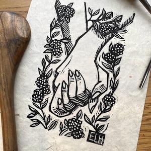 May include: Black and white linocut print featuring two hands holding each other, surrounded by floral elements and berries. The print is on textured paper, with the initials "ELH" in a small box. A wooden tool is visible on the left.