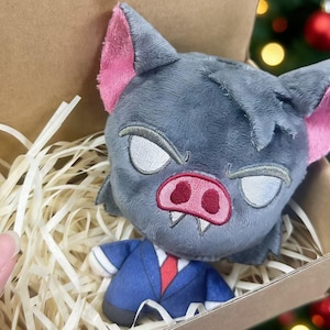 May include: A plush toy bat with gray fur, pink ears, and a red nose, presented in a cardboard box with paper filler. The bat has an angry expression and is wearing a blue suit with a red tie. The background is blurred.