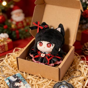 Puede incluir: Una muñeca de peluche negra con ojos rojos y un vestido rojo y negro está en una caja de cartón. La muñeca tiene orejas de gato y un lazo rojo. También son visibles dos llaveros, uno con una ilustración de personaje y otro con un emblema azul y dorado.