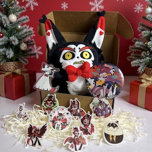 May include: A plush toy with a black and white face, red bow tie, and heart-shaped accents, sits in an open cardboard box. The box is surrounded by stickers, a button, and a clear keychain. The background includes Christmas trees and wrapped gifts.