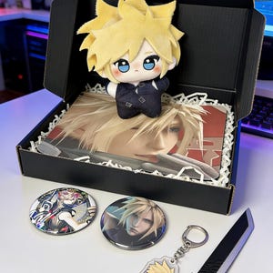 May include: A black gift box containing a plush doll with yellow hair, two round badges, a keyring, and a black sword replica. The doll has blue eyes and is wearing a dark outfit. The badges and keyring feature character art.
