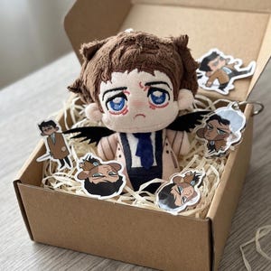 Op de afbeelding: Een pluche pop met bruin haar, blauwe ogen en zwarte vleugels, gekleed in een beige jas en blauwe das, in een kartonnen doos met vier kleine stickers en een doorzichtige acryl sleutelhanger met een cartoonfiguur in een bruine jas.