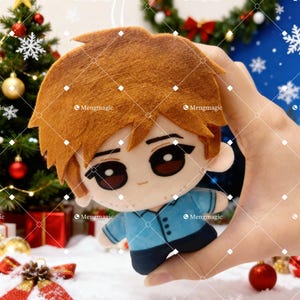 May include: A plush doll ornament featuring a cartoon character with brown hair and large eyes. The doll wears a blue shirt and black pants. The background includes a Christmas tree, ornaments, and gift boxes, suggesting a holiday theme.