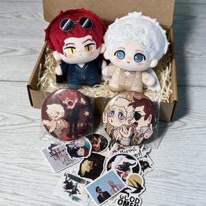 May include: Two plush dolls with large eyes, one with red hair and black sunglasses, the other with white hair. Included are two circular pin-back buttons featuring illustrated characters, and a collection of various small stickers in a box.