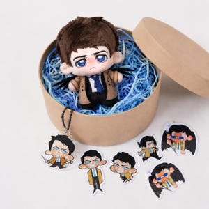 May include: A plush doll of a character with brown hair, blue eyes, and a tan coat, inside a round cardboard box with blue paper filler. Several character-themed acrylic charms are also present, featuring the same character in various poses.