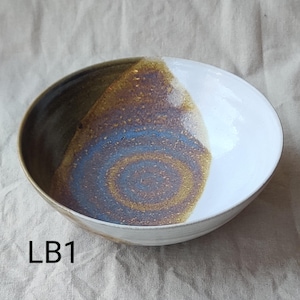 May include: A ceramic bowl with a distinctive design. The bowl has a white section and a section with brown, blue, and gold tones. The text "LB1" is visible on the bottom left of the bowl. The bowl is likely handmade.