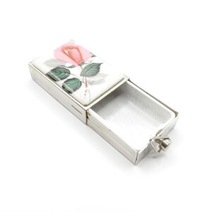 Peut inclure: Une boîte à allumettes en argent avec un couvercle à charnière. Le couvercle présente un motif peint d'une rose rose avec des feuilles vertes sur fond blanc.