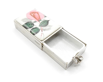 Puede incluir: Una caja de cerillas de plata con una tapa con bisagras. La tapa presenta un diseño pintado de una rosa rosa con hojas verdes sobre un fondo blanco.