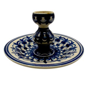May include: A blue and white ceramic candle holder. The candle holder has a circular base with a central raised section and a small, cup-shaped top. The design features detailed blue floral patterns on a white background, typical of ceramic art.
