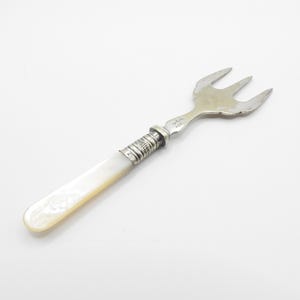 Könnte beinhalten: Eine Silberforke mit drei Zinken und einem Griff aus Perlmutt. Der Griff ist mit einem dekorativen Silberband an der Gabel befestigt. Die Gabel dient wahrscheinlich zum Servieren oder Essen einer kleinen Portion.
