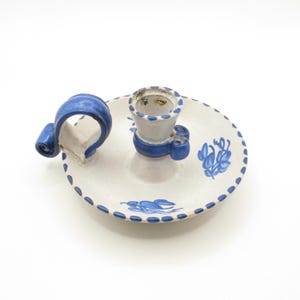 May include: A white ceramic candle holder with a blue handle and blue floral design. The holder has a round base and a raised center for the candle.