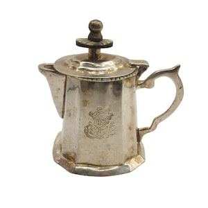Puede incluir: Una pequeña cafetera plateada con tapa y asa. La cafetera tiene un diseño ornamentado en la parte delantera y un adorno decorativo en la tapa. La cafetera probablemente está hecha de metal y está diseñada para servir café o té.