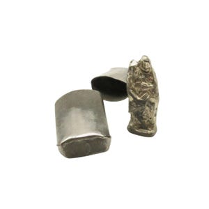 May include: Three silver-toned metal objects against a white backdrop. One is a small, open, cylindrical container. Another is a small, open, bell-shaped object. The third is a small, textured, abstract sculpture.