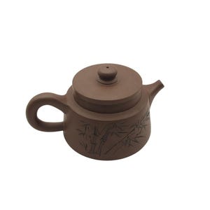 Puede incluir: Una tetera de cerámica marrón con tapa y asa. La tetera tiene un diseño de plantas de bambú grabado en el lateral.