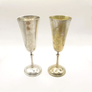 Puede incluir: Dos copas de champán vintage. Una es plateada, la otra dorada. Ambas tienen un tallo delgado y una copa cónica y ancha. Las copas están sobre un fondo blanco, resaltando su elegante diseño. Ideales para ocasiones especiales.