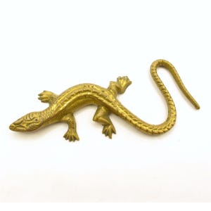 May include: A gold-toned brass lizard figurine featuring a textured, scaled body, four distinct legs with clawed feet, and a long, curved tail that arches upwards. The surface displays a metallic sheen with subtle variations in colour and finish.