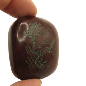 Puede incluir: Una piedra lisa de color marrón oscuro con un diseño de paisaje grabado en verde. El diseño incluye un árbol, un puente, montañas y otros elementos naturales. La piedra se sostiene entre dos dedos.