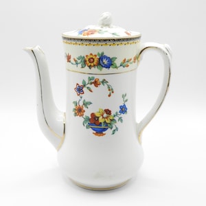 Op de afbeelding: Witte koffiekan van keramiek met een bloemenmotief in tinten oranje, geel, blauw en groen. De kan heeft een gouden rand en een deksel met een knop.