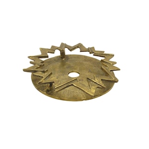 May include: A brass-colored, circular object with a central hole and a jagged, starburst-like design around the edge. A series of zig-zag shapes form a handle above the center. The object has a vintage or antique appearance.