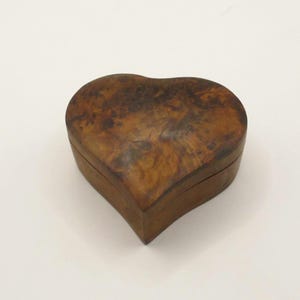 Può includere: Una scatola di legno a forma di cuore con finitura a venatura naturale. La scatola è di colore marrone caldo e ha un aspetto liscio e lucido. La scatola viene probabilmente utilizzata per riporre piccoli oggetti.