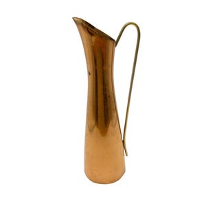 Peut inclure: Une cruche vintage en cuivre avec un corps élancé et allongé et une poignée en laiton. La cruche a un bec incurvé et une finition polie, reflétant la lumière. La poignée est de conception simple et incurvée. Idéal pour les fleurs ou comme décoration de cuisine.